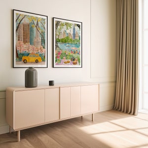 May include: Two framed watercolor paintings of New York City scenes. The painting on the left shows a yellow taxi cab driving through Central Park with skyscrapers in the background. The painting on the right shows a picnic scene in Central Park with a view of the city skyline.