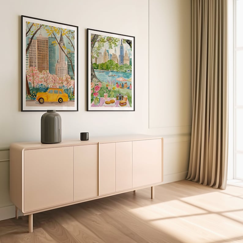 May include: Two framed watercolor paintings of New York City scenes. The painting on the left shows a yellow taxi cab driving through Central Park with skyscrapers in the background. The painting on the right shows a picnic scene in Central Park with a view of the city skyline.