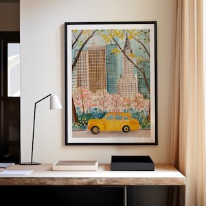 May include: A watercolor painting of a yellow taxi cab driving through Central Park in New York City. The Chrysler Building is visible in the background, and the trees are in bloom. The painting is framed in black.