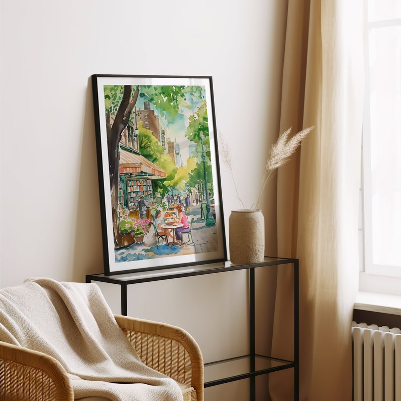 May include: A watercolor painting of a street scene in New York City. The painting depicts a bookstore with a cafe in front of it. People are walking and sitting at tables outside the cafe. The painting is framed in a black frame.