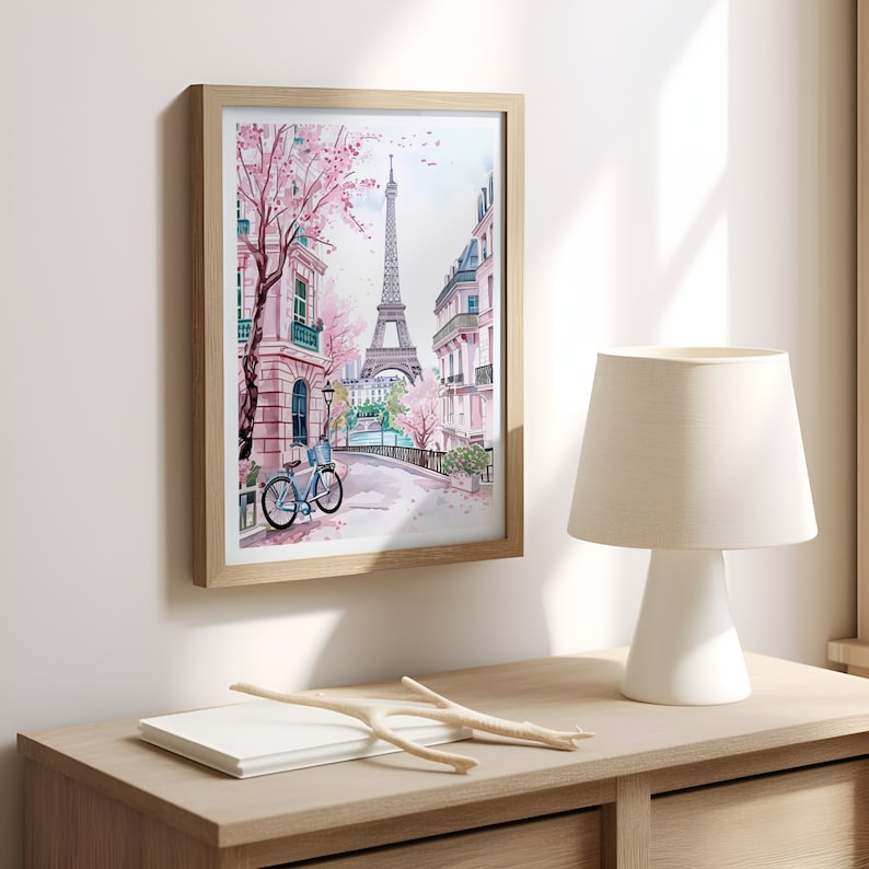 May include: A framed watercolor painting of a Parisian street scene with the Eiffel Tower in the background. The street is lined with pink buildings and a bicycle is parked on the side of the road. The painting is framed in a light wood frame.