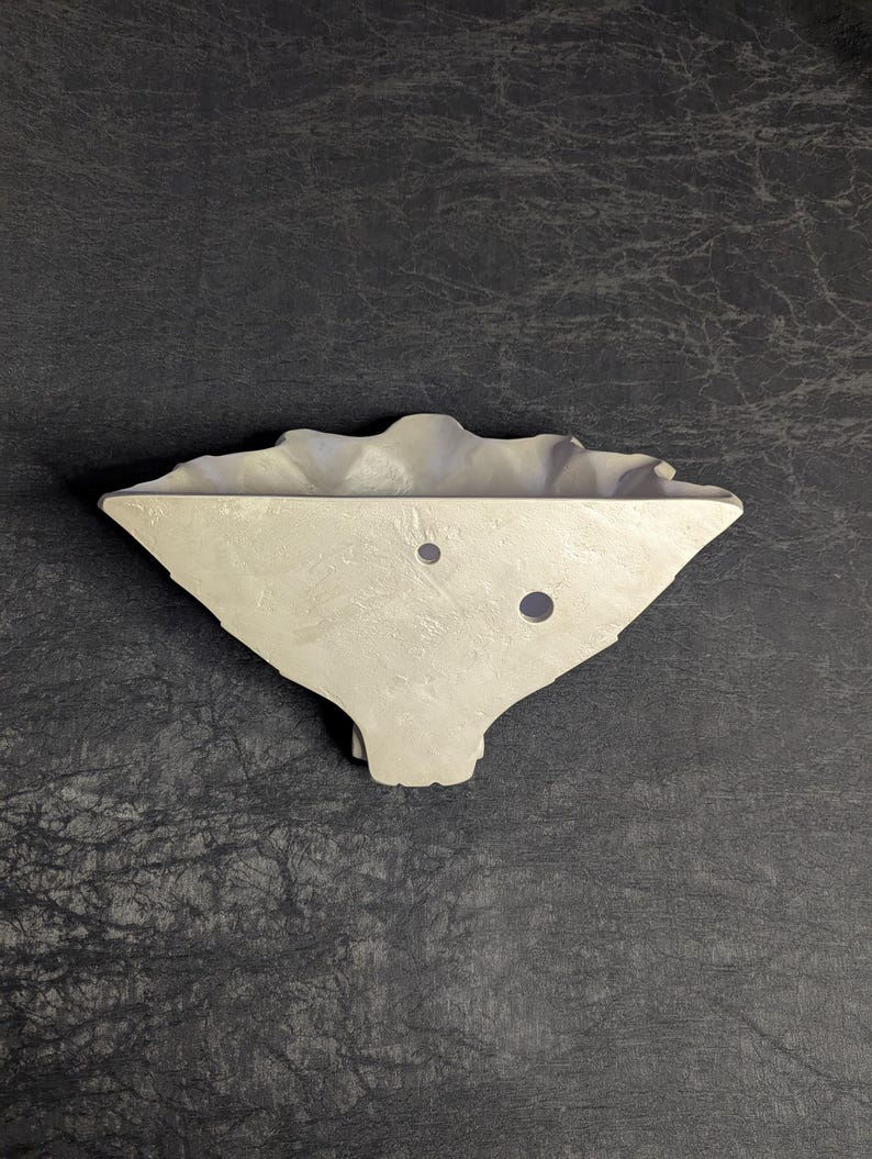 May include: A light gray, asymmetrical, wall-mounted planter with a textured surface. The planter has a wavy top edge and two circular holes. The design is modern and minimalist, suitable for small plants or succulents.