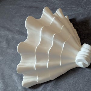 May include: A white, sculptural object with a fan-like design. The piece features a series of curved, wave-like ridges that create a textured surface. The object has a rounded, cylindrical base. The overall aesthetic is modern and minimalist.