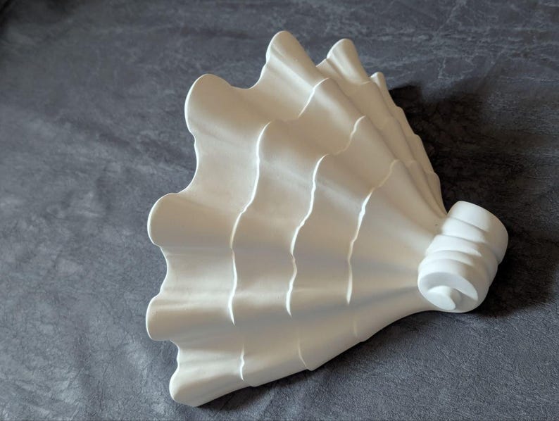 May include: A white, sculptural object with a fan-like design. The piece features a series of curved, wave-like ridges that create a textured surface. The object has a rounded, cylindrical base. The overall aesthetic is modern and minimalist.