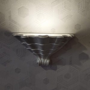 May include: A decorative, silver-colored wall shelf with a scalloped edge and a flared, textured base. The shelf is mounted on a gray wall with a geometric hexagonal pattern. The shelf is illuminated by a spotlight.