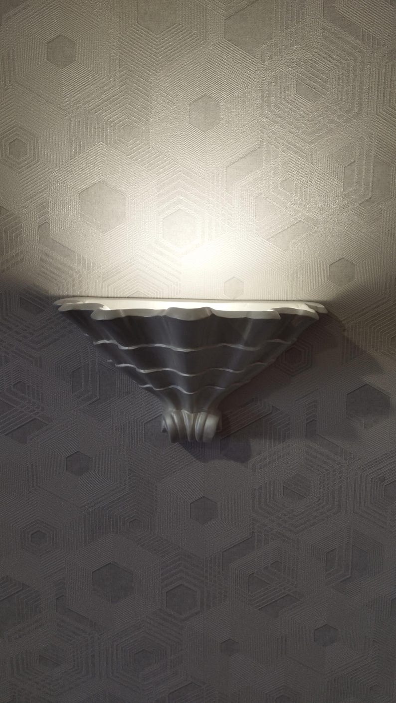 May include: A decorative, silver-colored wall shelf with a scalloped edge and a flared, textured base. The shelf is mounted on a gray wall with a geometric hexagonal pattern. The shelf is illuminated by a spotlight.