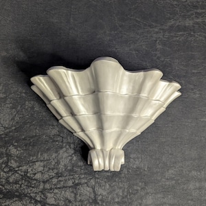 May include: A white, seashell-shaped wall sconce. The sconce has a detailed, ridged design, with a fan-like shape. The base of the sconce has a series of vertical, curved lines. The background is a dark, textured surface.