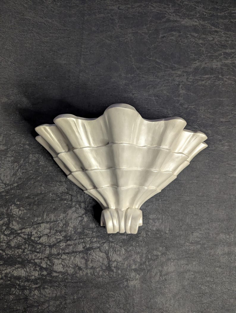 May include: A white, seashell-shaped wall sconce. The sconce has a detailed, ridged design, with a fan-like shape. The base of the sconce has a series of vertical, curved lines. The background is a dark, textured surface.