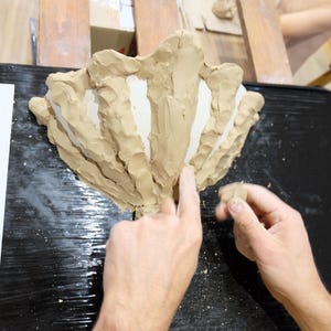 May include: A person's hands working on a clay sculpture of a stylized flower or shell. The sculpture is light brown and sits on a black surface. A sketch of a flower is on a white paper.