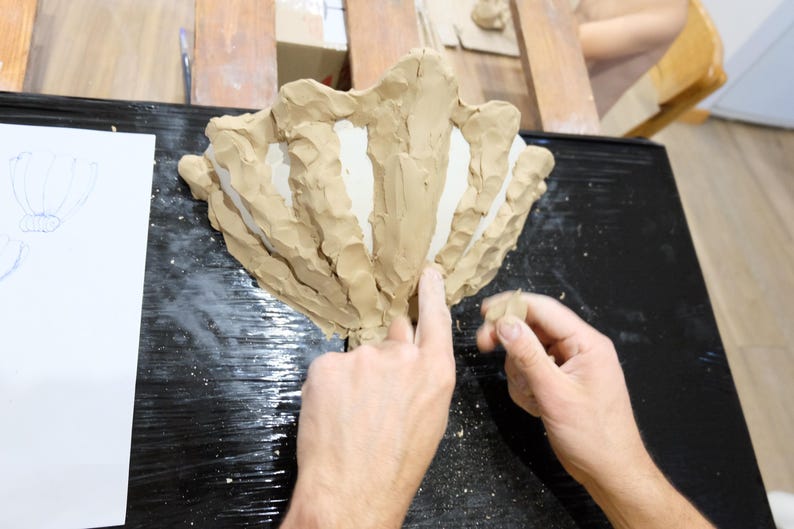 May include: A person's hands working on a clay sculpture of a stylized flower or shell. The sculpture is light brown and sits on a black surface. A sketch of a flower is on a white paper.