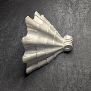 May include: A white, decorative architectural element with a fan-like design. The piece has a series of curved ridges and a cylindrical base with a circular detail. The object is set against a dark, textured background.