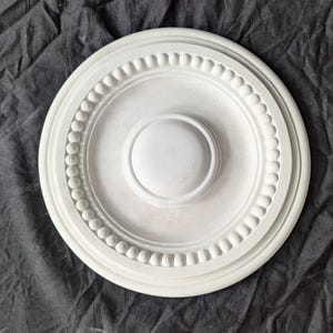 May include: A white, circular ceiling rose featuring a central raised disc and a beaded outer edge. The decorative plasterwork is designed for interior use. The design is symmetrical and classic, suitable for period properties.