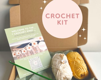 Op de afbeelding: Een haakpakket in een kartonnen doos, met garen, een haaknaald en instructies. De kit bevat twee garenbollen, één crème en één gele. De instructies luiden "Welkom bij de Crochet Kind!" en "Haakpakket".
