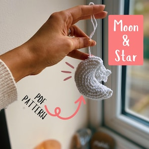 May include: A white crocheted moon and star ornament with a string attached. The ornament is being held by a hand. The text "Moon & Star" and "PDF PATTERN" are written on the image.
