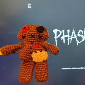 May include: A handmade brown crochet doll with black stitched features, a red eye, and a yellow patch. The word "PHASMOPHOBIA" is displayed in white, stylized text on a dark background.
