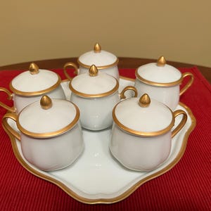 May include: A white porcelain tea set with gold trim, comprising six lidded cups and a matching tray. The cups feature curved handles and a rounded shape. The set is arranged on a red surface.