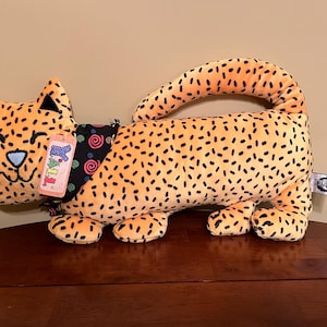 Puede incluir: Un gato de peluche naranja con motas negras, en posición tumbada. Tiene un collar negro y azul y una nariz azul. La etiqueta dice "Y'ALL". El juguete está sobre una superficie de madera.
