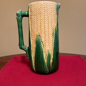 Puede incluir: Una jarra alta de cerámica con un diseño de mazorca de maíz. La jarra presenta un asa verde y hojas verdes, con un patrón de maíz amarillo y blanco. La jarra está sobre un mantel individual rojo.