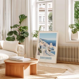 May include: A framed print of a colorful illustration of Santorini, Greece. The print features a white building with blue accents and a winding path leading to the ocean. The text "SANTORINI" is printed in blue at the bottom of the print.
