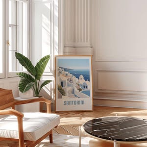 May include: A framed print of a colorful illustration of Santorini, Greece. The print features white buildings with blue accents, a winding street, and a view of the ocean. The text "SANTORINI" is printed at the bottom of the print.