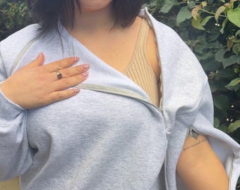 May include: A light gray sweatshirt with a zipper that extends around the shoulders. The sweatshirt is worn over a beige tank top and a beige skirt. The model is wearing a ring and has a tattoo on their arm.