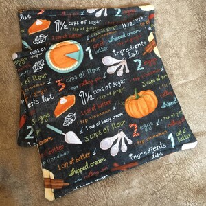 Puede incluir: Dos agarraderas de olla de tela negra y blanca con un diseño de pizarra que presenta una receta de pastel de calabaza. La receta incluye ingredientes como azúcar, mantequilla, huevos, harina, canela y jengibre. Las agarraderas están decoradas con imágenes de calabazas, pastel y cucharas medidoras.