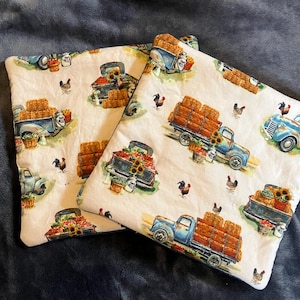 Könnte beinhalten: Zwei weiße Baumwoll-Topflappen mit einem blauen LKW-Design. Die LKWs transportieren Heuballen und Sonnenblumen. Der Hintergrund ist hellcremefarben mit einem Muster aus Hühnern und Sonnenblumen.