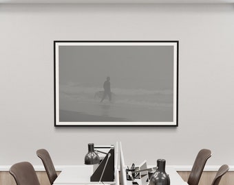 May include: A black framed photograph of a person walking on a beach with a surfboard. The photo is hanging on a white wall in an office setting. There are two desks with chairs and lamps in the foreground.