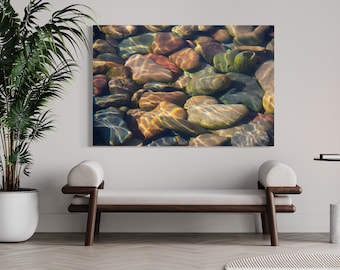 May include: A close-up photo of smooth, colorful river rocks submerged in clear water. The sunlight shines through the water, creating a shimmering effect on the rocks.