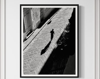 May include: A black and white photograph of a person walking down a cobblestone street in a city. The person is walking towards the camera and their shadow is cast long on the ground. The street is lined with buildings on either side.