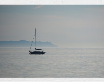 May include: A white sailboat with a blue sail is anchored in a calm, misty body of water. The water is a light blue color and the sky is a pale blue. There are mountains in the distance.