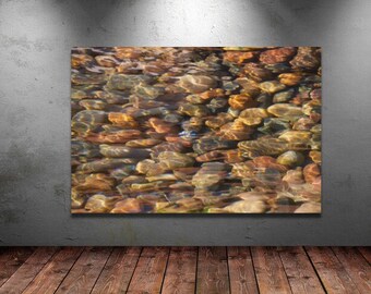 May include: A close-up image of smooth, colorful river rocks submerged in clear, shallow water. The rocks are various shades of brown, red, and gray, with the water reflecting light and creating a shimmering effect.