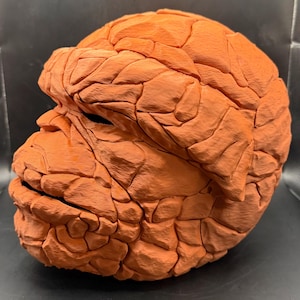 Pode incluir: Uma m&aacute;scara laranja, texturizada, que se assemelha a uma personagem de banda desenhada. A m&aacute;scara &eacute; de cor s&oacute;lida com uma textura rugosa, semelhante &agrave; pedra, e apresenta uma testa, nariz e queixo proeminentes. Provavelmente uma pe&ccedil;a de fantasia ou colecion&aacute;vel.