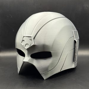 May include: A silver-colored helmet with a stylized design. The helmet features a prominent crest with a geometric emblem above the eye openings. The design includes ridges and a textured surface, suggesting a futuristic or superhero theme.