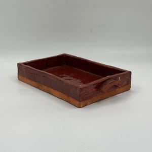 Può includere: Una pirofila rettangolare in terracotta con interno e bordo smaltati in marrone scuro lucido. I lati esterni presentano una finitura in argilla naturale non smaltata di colore beige chiaro. Due piccole maniglie curve sui lati corti.