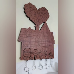 Può includere: Un portachiavi in legno a forma di casa con un gruppo di palloncini a forma di cuore in cima. La casa ha cinque personaggi del film Up, tra cui Carl ed Ellie. Ci sono cinque ganci per le chiavi.