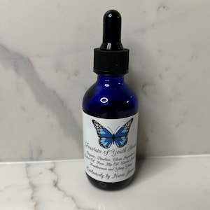 Puede incluir: Una botella de vidrio azul cobalto con un cuentagotas negro. La etiqueta presenta una mariposa azul y el texto "Fountain of Youth Serum". La botella contiene ingredientes orgánicos y es exclusivamente de Nurse Jenna.