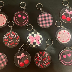 Könnte beinhalten: Eine Sammlung von Acryl-Schlüsselanhängern mit verschiedenen Designs. Die Schlüsselanhänger zeigen rosa und schwarze Karomuster, Kirschen, Schleifen und Smiley-Gesichter mit Herzen. Jeder Schlüsselanhänger hat einen silbernen Metallring.