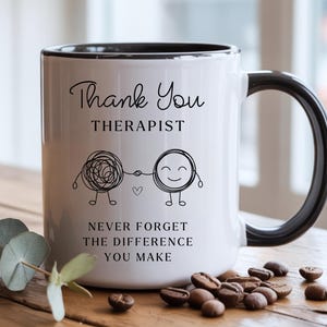 Könnte beinhalten: Weißer Keramikbecher mit schwarzem Innenraum und Henkel. Der Becher trägt den Text "Thank You THERAPIST" und den Satz "NEVER FORGET THE DIFFERENCE YOU MAKE". Eine skurrile Illustration von zwei verbundenen Figuren ist ebenfalls vorhanden.