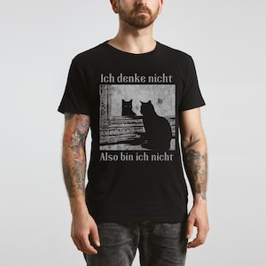 May include: Black t-shirt featuring a graphic of a cat looking in a mirror at its reflection. The mirror image and the cat's silhouette are in black and white. The text "Ich denke nicht, Also bin ich nicht" is above and below the image.