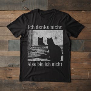 May include: Black t-shirt with a graphic of a cat looking in a mirror at its reflection. The mirror image is also a cat. The text on the shirt reads "Ich denke nicht, Also bin ich nicht."