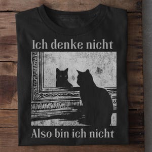 May include: Black t-shirt featuring a graphic of a cat looking in a mirror at its reflection. The mirror image and the cat are black silhouettes. The text "Ich denke nicht, Also bin ich nicht" is printed in white.