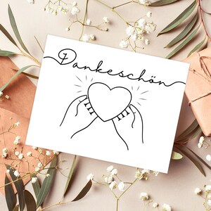 Può includere: Un biglietto bianco con inchiostro nero che dice "Dankeschön" in corsivo. Un cuore è disegnato al centro del biglietto con due mani che lo tengono. Il cuore ha un bagliore raggiante intorno a sé.
