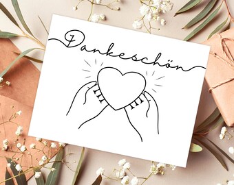 May include: A white card with black ink that says "Dankeschön" in cursive. A heart is drawn in the center of the card with two hands holding it. The heart has a radiant glow around it.