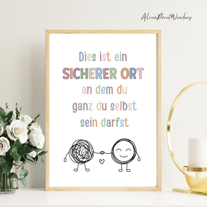 May include: A framed print with the text "Dies ist ein SICHERER ORT an dem du ganz du selbst sein darfst" in pastel colors. Below the text are two cartoon figures holding hands, one a tangled scribble, the other smiling. The print is in a light wood frame.