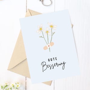 May include: A light blue card with a white flower bouquet and a bandage with a red heart. The card says "Gute Besserung" in black text.