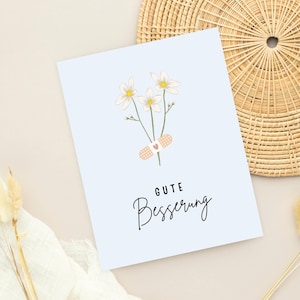 May include: A light blue card with a white flower bouquet and a bandage with a red heart. The card says "Gute Besserung" which is German for "Get well soon".