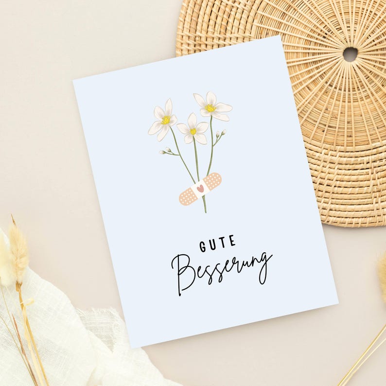 May include: A light blue card with a white flower bouquet and a bandage with a red heart. The card says "Gute Besserung" which is German for "Get well soon".
