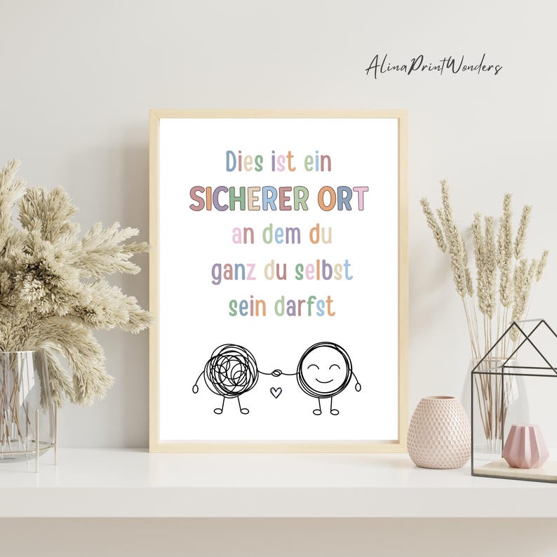 May include: A framed print with the text "Dies ist ein SICHERER ORT an dem du ganz du selbst sein darfst" in pastel colors. The print features two cartoon figures holding hands, one with a tangled design and the other with a smiling face.