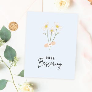 May include: A light blue card with a white flower bouquet and a bandage with a heart on it. The card says "Gute Besserung" which is German for "Get well soon".
